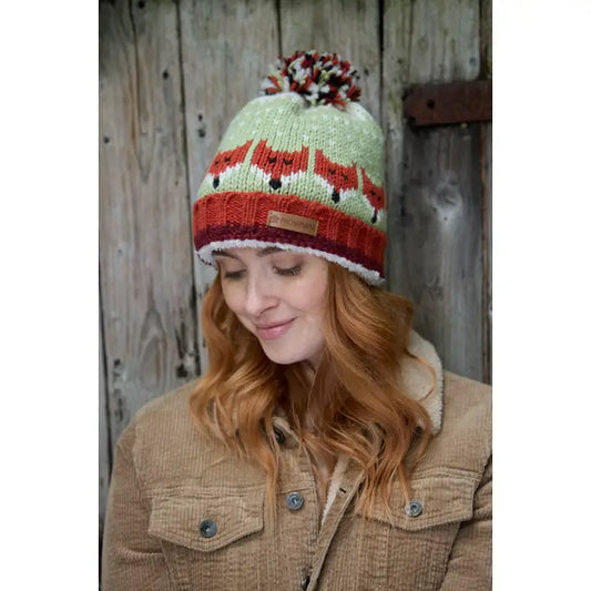 Frau trägt Skulk of Foxes Fair Isle Wollmütze mit Fuchsmotiv und Bommel – handgestrickt mit Fleecefutter
